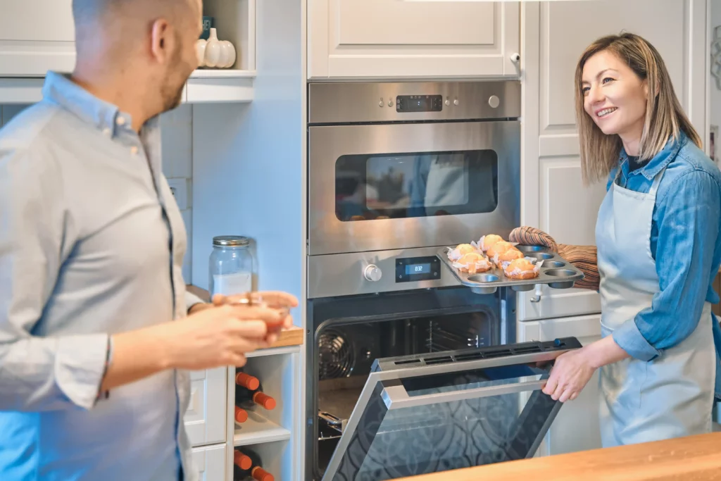 A woman takes a tray of muffins out of an oven while smiling at a man holding a cup in a modern kitchen.