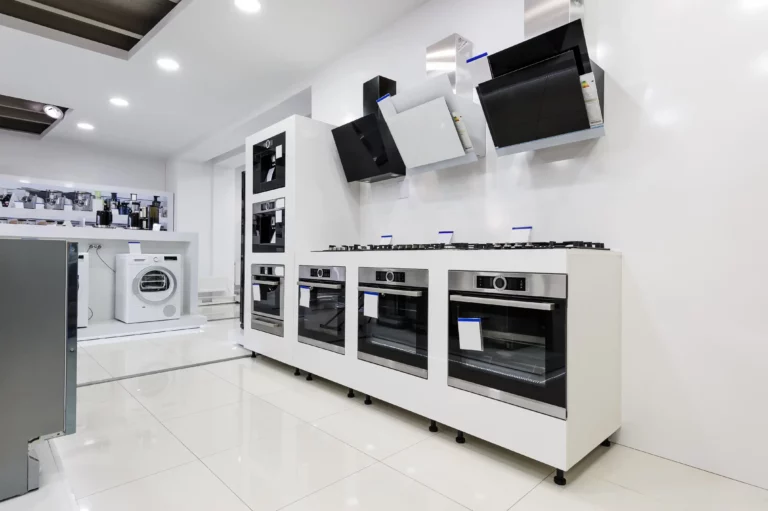 An elite showroom displays modern built-in ovens, stovetops, and range hoods along a white wall, with a washing machine visible in the background.