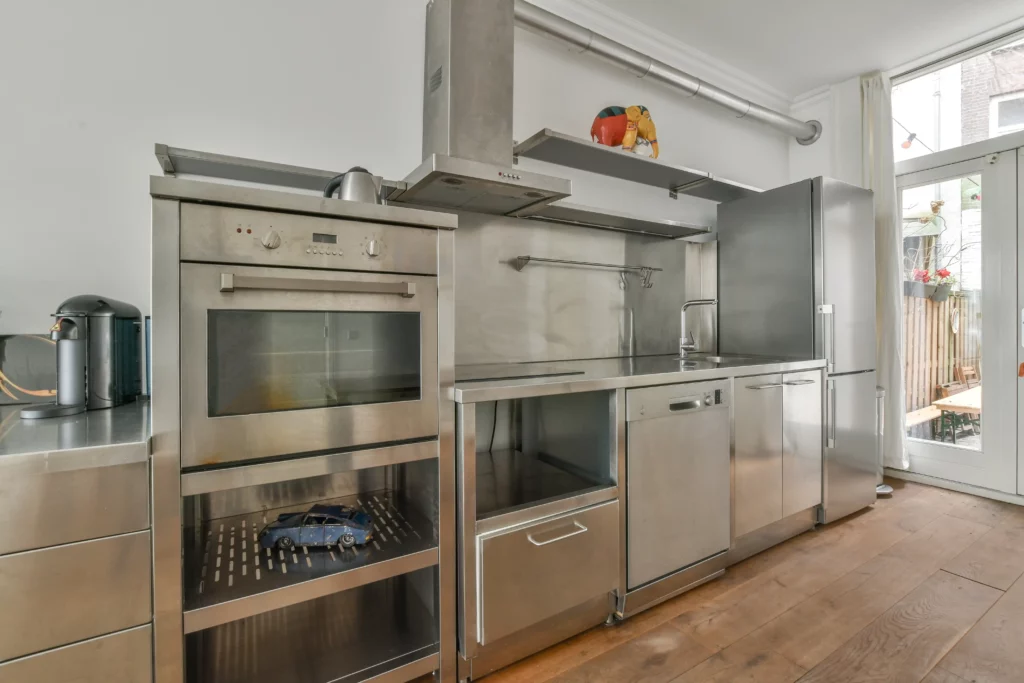 Modern kitchen with stainless steel appliances, including an oven, dishwasher, sink, and refrigerator, set against a white wall with wooden flooring and large glass doors.