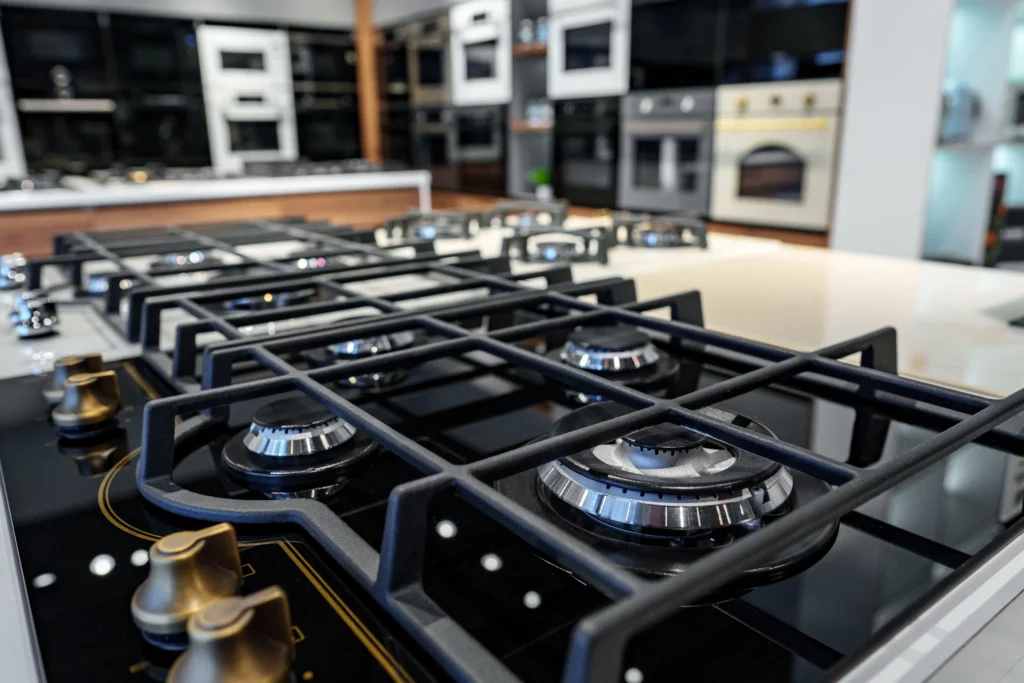 Close-up of a modern gas stove cooktop with four burners and metallic control knobs in a kitchen appliance showroom. Other ovens and stoves are visible in the background.