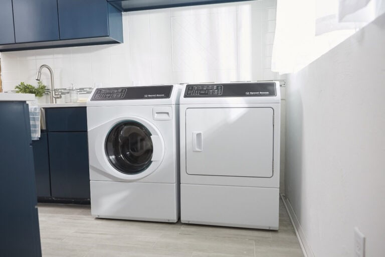 A modern laundry room with a front-loading washing machine and a matching dryer side by side, blue cabinets, and a window letting in natural light.