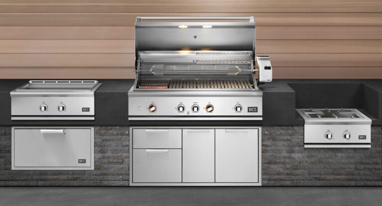A modern outdoor kitchen setup featuring a large stainless steel grill with open lid, side burners, storage drawers, and cabinets against a stone and wood backdrop.
